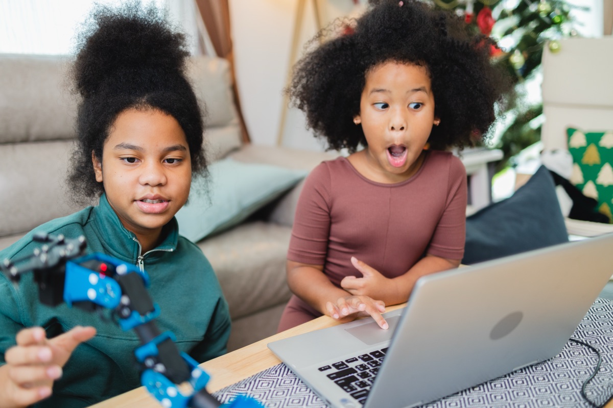 Two girls at a laptop with a robotic arm, one reacting with delight